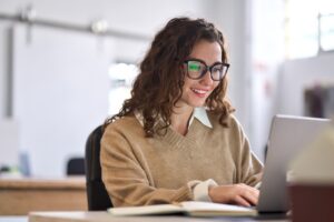 A woman with glasses and long, curly brown hair sitting in front of a computer with a smile on her face. | Call center sales blog post