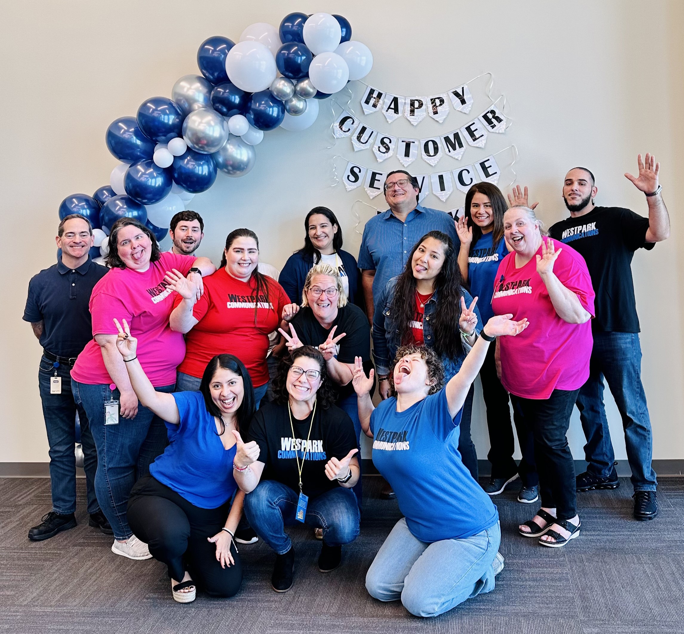 group of employees dressed in blue, pink, and red shirts with balloons in front of a sign that says "Happy Customer Service Week"